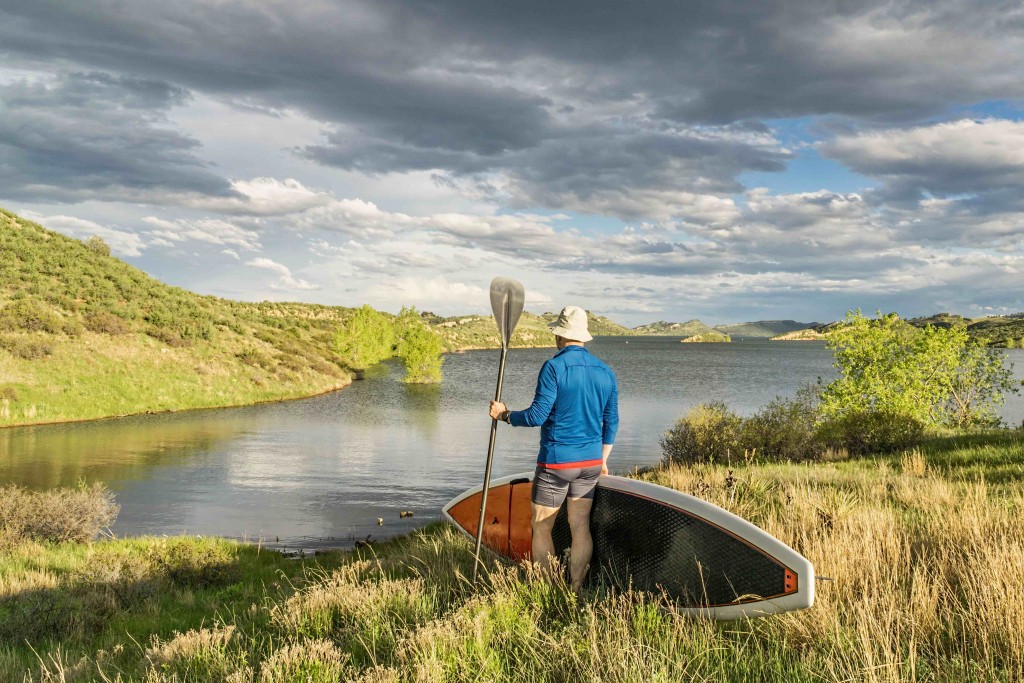 male paddler with stand up paddleboard (SUP) on a grassy shore of mountain lake - Horsetooth Reservoir near Fort Collins, Colorado